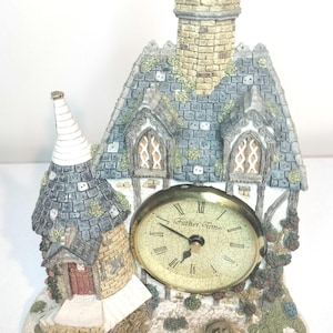 May include: A decorative table clock shaped like a cottage with a stone chimney, a conical tower, and a textured roof. The clock face is oval with a cracked cream background, black Roman numerals, and the text Father Time printed in black script.