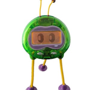 May include: A green, alien-shaped electronic toy with a digital display. It has yellow legs and antennae with red tips. The display shows two oval shapes. The toy has a purple frame around the display and purple feet.