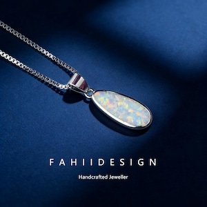May include: A silver necklace with a teardrop-shaped pendant. The pendant has a white opal with iridescent colours. The necklace is on a dark blue background. The text "FAHIIDESIGN" and "Handcrafted Jeweller" are at the bottom.