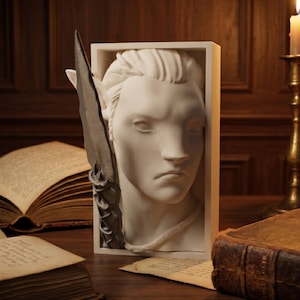 Puede incluir: Un busto blanco de una persona con orejas puntiagudas, enmarcado en un rectángulo. Una pluma o cuchilla oscura y estilizada está colocada a la izquierda. El busto está sobre una superficie de madera, con libros abiertos y una vela encendida en el fondo.