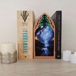 Puede incluir: Un inserto de rincón de libro arqueado en color marrón claro que presenta una escena forestal detallada e iluminada con un arroyo, linternas brillantes y ramas de árboles con hojas verdes. El marco puntiagudo se sitúa entre dos libros.