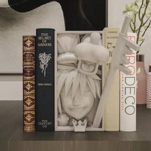 Può includere: Un'esposizione artistica con libri e oggetti decorativi. Una scultura bianca scolpita di un personaggio è incorniciata da una scatola bianca. Sono visibili libri con titoli come "The Helmet of Navarre" e "Art Deco", insieme a vasi e una scultura decorativa.