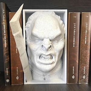 May include: A light gray speckled bookend featuring a sculpted, snarling humanoid face with prominent brow ridges and sharp teeth, housed within a rectangular frame, positioned between several brown books with visible text on their spines.