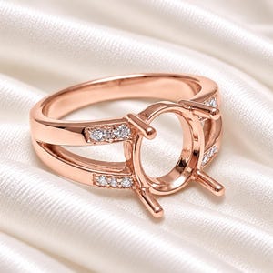 May include: A rose gold ring setting featuring an oval-shaped center mount with four prongs. The split shank design is accented with small, clear round stones embedded along the sides of the band, creating a polished and metallic appearance.