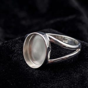 Peut inclure: Une bague en argent avec un sertissage de lunette ovale. La bague a un design à tige fendue, avec deux bandes parallèles incurvées autour du doigt. Le sertissage est conçu pour contenir une pierre précieuse. La bague est présentée sur une surface noire.