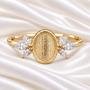 Könnte beinhalten: Ein goldener Ring mit einer zentralen, ovalen Fassung mit texturierter, matter Oberfläche. Der Ring ist auf beiden Seiten mit drei klaren, marquise-geschliffenen Steinen in blattartiger Anordnung verziert, was ein symmetrisches Design ergibt.