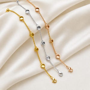 Könnte beinhalten: Drei filigrane Armbänder in Gold, Silber und Roségold. Jedes Armband hat eine Kette mit gleichmäßig verteilten, kreisförmigen Fassungen. Präsentiert auf weißem, seidigem Stoff, der die verschiedenen Metalltöne hervorhebt.