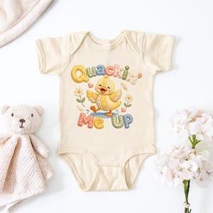 May include: A cream-colored baby onesie with a colorful graphic of a yellow duckling with the text "Quackin' Me Up". The onesie is surrounded by flowers and is next to a teddy bear and a blanket.