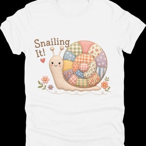 May include: White t-shirt featuring a whimsical snail design. The snail has a patchwork shell in various pastel colors and the text "Snailing It!" above it. The design includes flowers and a heart.