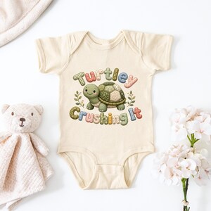 May include: A cream-colored baby onesie with a cartoon turtle graphic. The turtle has a green shell and is surrounded by the words "Turtley Crushing It" in colorful, patterned letters. A teddy bear and flowers are in the background.