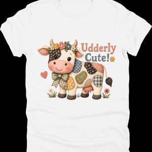 May include: White t-shirt with a cartoon cow design. The cow has a patchwork pattern, a floral crown, and a bow. The text "Udderly Cute!" is printed in red and orange. The design includes a heart and flowers.