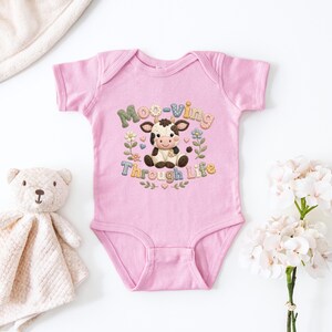 May include: A pink baby onesie with a cartoon cow design and the words "Moo-ving Through Life". The design includes flowers and hearts. A stuffed teddy bear and a blanket are in the background.