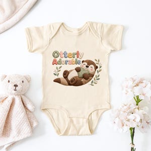 Könnte beinhalten: Ein cremefarbener Baby-Body mit dem Text "Otterly Adorable" über einer Aquarellillustration eines braunen Otters. Der Otter liegt und hält eine grüne Decke. Ein Teddybär und Blumen sind ebenfalls im Bild zu sehen.