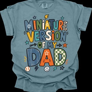May include: A blue t-shirt with the text "MINIATURE VERSION OF MY DAD" in colorful, playful lettering. The design includes stars, lightning bolts, a baseball, and a smiley face. The shirt is made of a soft material.
