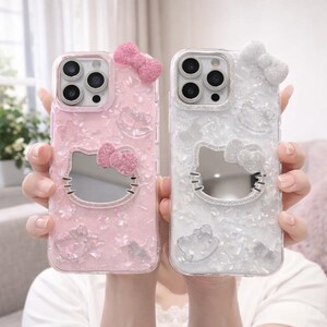 May include: Two phone cases, one pink and one white, featuring a Hello Kitty design. Each case has a bow at the top and a mirrored Hello Kitty face. The pink case has pink glitter, and the white case has white glitter.