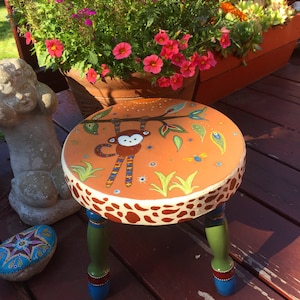 May include: A hand-painted, round wooden stool with a whimsical monkey design. The seat is orange with a monkey hanging from a branch, surrounded by leaves and insects. The edges have a giraffe-like pattern, and the legs are green, blue, and red.