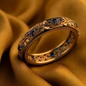 Puede incluir: Un anillo de banda de oro con un intrincado patrón en relieve de hojas y enredaderas, acentuado con pequeñas piedras preciosas redondas de color azul profundo engastadas a intervalos en la superficie exterior y en el diseño calado.