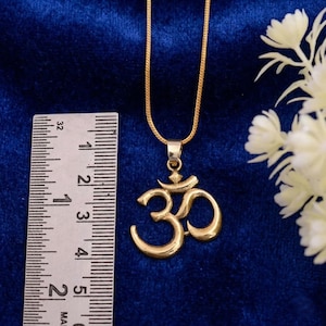 Puede incluir: Un collar con colgante Om dorado. El colgante mide aproximadamente 4 cm de altura y presenta el símbolo Om. El collar se muestra sobre un fondo azul con una regla para la escala.