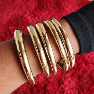 Op de afbeelding: Een set van vijf gepolijste goudkleurige metalen armbanden gedragen om de pols. Elke armband heeft een glad, afgerond en doorlopend cirkelvormig ontwerp met een hoogglanzend reflecterend oppervlak dat het licht vangt voor een uniforme uitstraling.