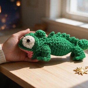 May include: A green crocheted lizard toy featuring a textured body, a prominent white eye patch with a black pupil, and a pink mouth area. The stuffed animal has a ridged back and four small legs, crafted with visible yarn stitches.