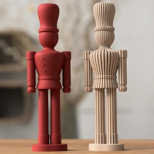 Könnte beinhalten: Zwei stehende Soldatenfiguren auf einem flachen Sockel. Die linke Figur ist einfarbig rot mit einer strukturierten Oberfläche. Die rechte Figur ist hellbeige und aus einer Reihe vertikaler Lamellen gefertigt, was ihr ein geripptes Aussehen verleiht.