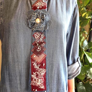 May include: A light blue denim shirt featuring a decorative necktie accessory. The tie is crafted from denim and deep red fabric, embellished with a denim flower, a central pearl, intricate floral embroidery, and scattered white beads.