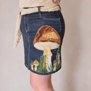 Peut inclure: Jupe en jean bleu foncé avec broderie de champignons. La jupe présente une coupe droite et une scène de champignons détaillée dans les tons marron, beige et vert. Le texte "Hand Embroidered Denim Skirt" est visible.
