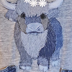May include: A denim patchwork design of a Highland cow with a white floral crown. The cow is made from various shades of blue denim, with black eyes and a light blue nose. The background is a heather gray fabric.