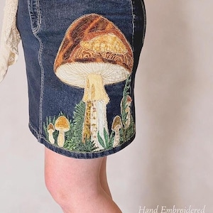 May include: A dark blue denim skirt featuring detailed hand embroidery of a large brown mushroom with a cream-colored stem, surrounded by smaller mushrooms and green foliage. The text "Hand Embroidered Denim Skirt" is visible in the bottom right corner.