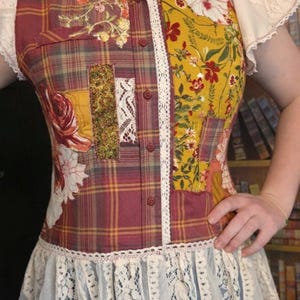 May include: A patchwork shirt with a plaid pattern in burgundy, yellow, and green. The shirt features floral and lace accents, with short lace sleeves and a lace trim at the bottom. The shirt has a button-down front.