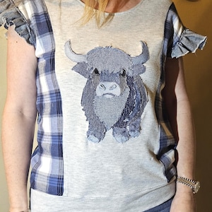 May include: A light gray short-sleeved top featuring a central applique of a shaggy highland cow made from denim fabric. The garment includes blue and white plaid side panels and ruffled sleeves, creating a textured look with a round neckline.