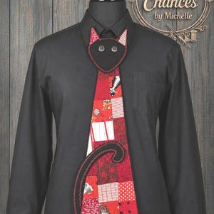 May include: A black long-sleeve button-down shirt worn with a novelty necktie featuring a patchwork design in shades of red, white, and black. The tie has a cat-shaped top with button eyes and a black cat tail appliqué on the front panel.