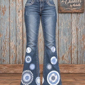 May include: Dark blue denim flare jeans featuring a high-waisted design with two silver buttons and white stitched detailing on the front pockets. The lower legs are decorated with various circular fabric patches in shades of blue and white.