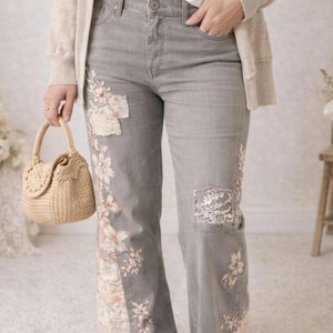 Könnte beinhalten: Graue Jeans mit floralen Stickereien und Patches. Die Jeans hat ein weites Bein und ist mit Spitzen- und Blumenmustern verziert. Eine kleine, gewebte Handtasche wird in der Hand gehalten.