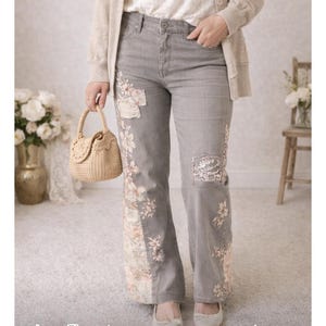 May include: Grey denim jeans with floral embroidery and patches. The jeans have a wide leg cut and are paired with a beige cardigan and a white lace top. A small woven handbag and ballet flats complete the outfit.