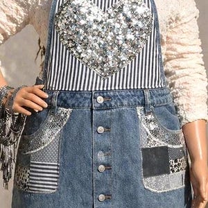 May include: A blue denim overall skirt featuring a central bib with a black and white vertical stripe pattern and a large silver sequined heart. The skirt has a button-down front and two front pockets decorated with mixed grey and black patches.