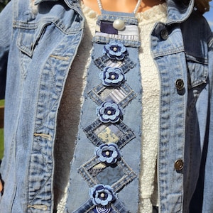 Könnte beinhalten: Eine Krawatte aus blauem Jeansstoff mit einem Patchwork-Design und ausgefransten Kanten. Die Krawatte ist mit mehreren geschichteten Stoffblumen in Blautönen verziert, die jeweils mit einer kleinen weißen Perle und einem weißen Knopf versehen sind.