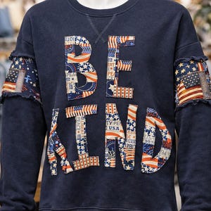 Può includere: Felpa blu navy con le parole "BE KIND" applicate in un design patchwork con la bandiera americana. Le maniche hanno un design a bandiera invecchiata. Uno stile casual e patriottico.