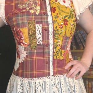 May include: A button-down shirt featuring a patchwork design of plaid fabric, mustard yellow floral panels, and rose-patterned sections. The garment includes white lace trim along the button placket, a ruffled lace hem, and short lace sleeves.