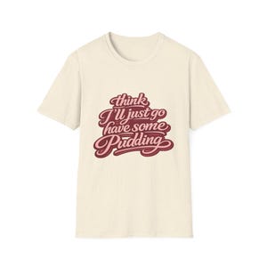 Puede incluir: Camiseta color crema con la frase "think I'll just go have some Pudding" en una fuente retro y cursiva. El texto está en tonos burdeos y rosa. La camiseta tiene cuello redondo clásico y mangas cortas.