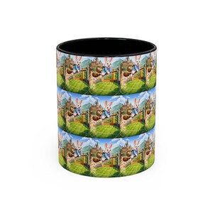 May include: A black-handled coffee mug with a repeating Easter-themed design. The design features a cartoon rabbit riding a broom over a green field, with a castle in the background. The rabbit is wearing a blue jacket and holding a basket of eggs.
