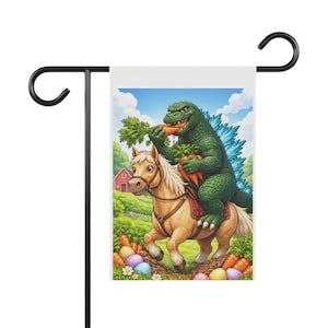 Puede incluir: Una bandera de jardín con una ilustración de dibujos animados de Godzilla montando un pony y comiendo zanahorias. El fondo incluye una escena de granja con un granero rojo, hierba verde y huevos de Pascua.
