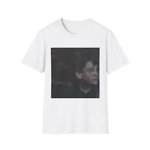 Könnte beinhalten: Weißes T-Shirt mit einem Porträt eines jungen Menschen mit runder Brille. Das Bild zeigt eine Nahaufnahme des Gesichts der Person mit einem nachdenklichen Ausdruck. Das T-Shirt besteht aus einem weichen, bequemen Material.