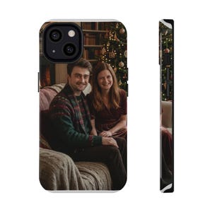 May include: A white iPhone case featuring a photograph of two people sitting on a couch in front of a Christmas tree. The case has a black camera lens and buttons on the side.