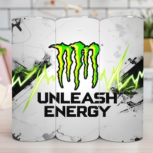 Könnte beinhalten: Ein weißer Becher mit einem Used-Design, das das Monster Energy-Logo in Grün und Gelb zeigt. Die Worte "UNLEASH ENERGY" sind darunter in Schwarz gedruckt, mit grünen Blitz-Akzenten.