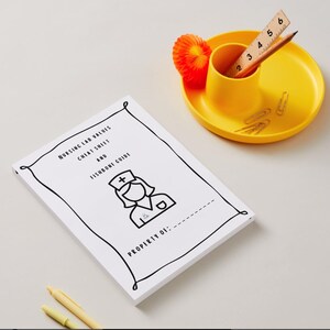 May include: A white booklet with the text "NURSING LAB VALUES CHEAT SHEET AND FISHBONE GUIDE" and a nurse illustration. A yellow tray holds a pencil, ruler, and paperclips. A small orange decorative item is also on the tray.