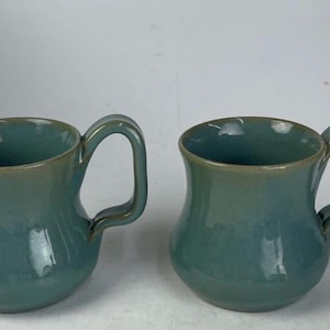 May include: A set of three ceramic mugs with a speckled, matte finish. The mugs are in shades of cream, light gray, and dark gray. Each mug has a simple, minimalist design and a rounded handle.