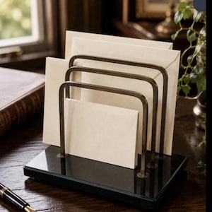 May include: A letter holder featuring three tiered, U-shaped antique brass metal dividers mounted on a rectangular, polished black stone base. The organizer holds several cream-colored envelopes of varying sizes, arranged in a cascading vertical display.