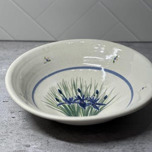 May include: A round, off-white ceramic bowl with a speckled finish. The interior features a hand-painted design of green grass and dark blue iris flowers at the center, encircled by a thin blue line, with small floral accents near the rim.