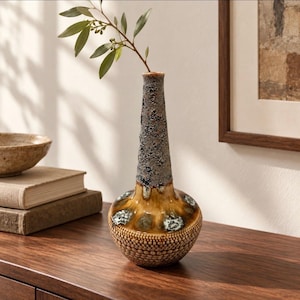 May include: A ceramic vase with a bulbous base and a tall, slender neck. The base features a textured, dotted pattern in brown, while the upper section has a rough, mottled gray and brown glaze with white accents holding a green leafy sprig.